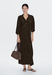 Bruine satijnen blouse met opgerolde mouwen en knoopsluiting, gecombineerd met een bijpassende lange rok. Bruine suède handtas en platte sandalen inbegrepen.