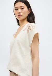 Νεαρή γυναίκα με μακριά σκούρα μαλλιά φοράει κρεμ τοπ με κοντά μανίκια, διακοσμημένο με κεντημένα καφέ και ροδακινί ανθισμένα σχέδια στους ώμους.