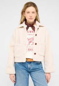 Cream-colored fleece jacket with a collared neck, two patch pockets, and brown buttons, paired with a graphic t-shirt and blue jeans.