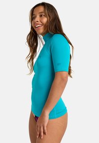 Krátky rukáv tyrkysovej ochranné tričko s vysokým golierom, hladkou textúrou a priliehavým strihom. Výrazný detail loga na rukáve.