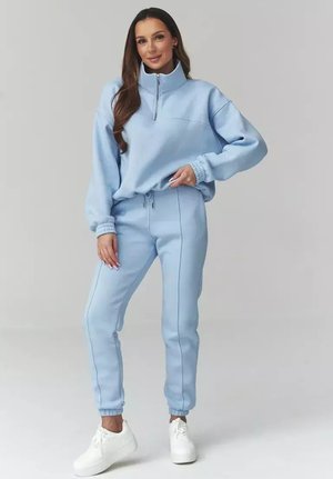 Kobieta w jasnoniebieskiej bluzie rozpinanej na zamek i pasujących spodniach dresowych, w białych trampkach, stoi z jedną ręką na biodrze na jednolitym tle.