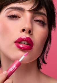 Brillo de labios rosa con acabado brillante aplicado con un aplicador pequeño desde un tubo a juego etiquetado como "Vamp! Marker Duo." El fondo es rosa.