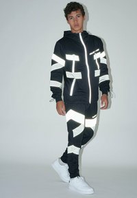 Chándal deportivo negro con acentos reflectantes, que incluye una chaqueta con cremallera y pantalones ajustados. Combinado con zapatillas blancas.