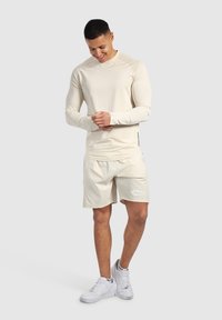 Maglietta atletica a manica lunga beige chiaro con tessuto texturizzato, abbinata a pantaloni corti coordinati con un logo bianco. Indossata con scarpe da ginnastica bianche.