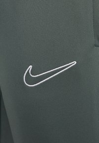Groene sportbroek met een opvallend wit geborduurd Nike-logo op de dij, gemaakt van een gladde, rekbare stof.