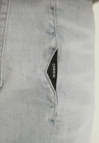 Jeans gris claro con una tela texturizada, con una distintiva abertura de bolsillo diagonal que muestra una etiqueta negra con la inscripción "CHASIN'".
