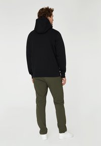 Sudadera negra con capucha grande, puños y bajo acanalados, combinada con pantalones verde olivo y zapatillas blancas, vista desde la parte trasera.