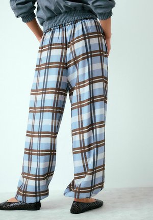 Osoba w luźnych, jasnoniebieskich, kratkowanych spodniach w brązowe i białe paski, czarne wsuwane buty, stojąca z rękami na biodrach.