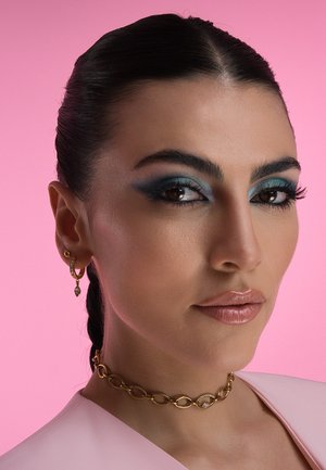Primo piano di un volto con capelli scuri. Caratterizzato da un ombretto blu audace e labbra lucide di nude. Collana choker in oro e orecchini con dettagli in diamante. Sfondo rosa.