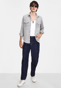 Camicia a bottoni grigia a fantasia sopra una canotta bianca, abbinata a pantaloni cargo blu scuro e sneakers bianche. Presenta occhiali da sole e gioielli in argento.