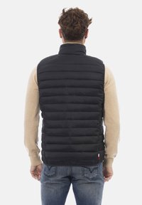 Schwarze Daunenweste mit hohem Kragen, horizontaler Steppung und Seitentaschen. Getragen über einem beigen Pullover mit hellblauen Jeans.