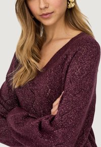 Maglione bordeaux con scollatura a V, maniche lunghe e tessuto texturizzato e lucido. Presenta una vestibilità ampia e polsini a costine.