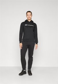 Svart hettegenser med hvit "Champion" logo, frontlomme, snorer; sammen med matchende svarte joggebukser med logo. Svarte sneakers.
