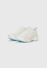 Ein Paar weiße Sportschuhe mit strukturierten Sohlen, subtilen Wellenmustern und blauen Gummisohlenakzenten, vor einem neutralen Hintergrund abgebildet.