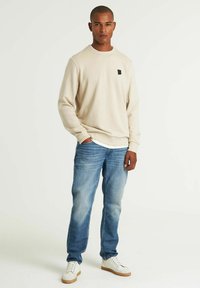 Sudadera beige de cuello redondo con un pequeño logo negro, combinada con jeans de mezclilla azul claro y zapatillas blancas con suelas de color marrón claro.