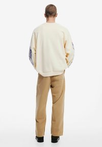 Crèmekleurige sweatshirt met lange mouwen, voorzien van grafische ontwerpen op de mouwen. Gepaard met lichtbruine broeken en zwarte schoenen.