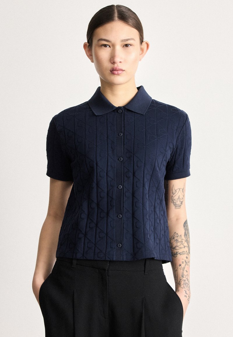 Tumši zila adīta polo krekls ar īsām piedurknēm, raksturīga teksturēta dimanta raksta, priekšējām pogām un klasisku apkakli.