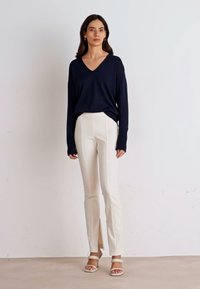 Marineblauwe V-hals longsleeve top, gecombineerd met witte op maat gemaakte broek met zijsplitten. Model draagt beige sandaaltjes met bandjes. Effen achtergrond.