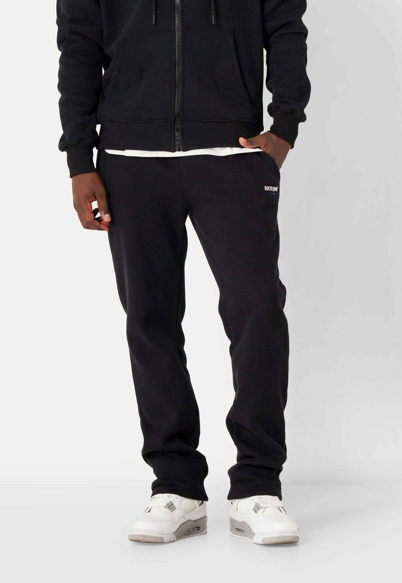 Schwarze Zip-Hoodie mit Fronttaschen, kombiniert mit schwarzen Jogginghosen mit einem Logo. Getragen mit weißen Sneakers. Material aus Baumwollmischung.