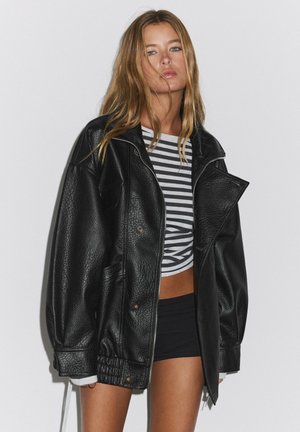 Jeune femme aux longs cheveux blonds portant une veste en cuir noire oversize, un short noir et un crop top rayé noir et blanc.