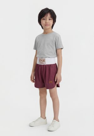 Garçon debout portant un t-shirt gris, un short de sport bordeaux avec une large ceinture blanche, et des baskets blanches sur un fond uni.