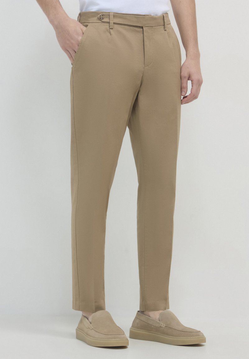 Mann in schmal geschnittenen beigen Hosen mit seitlichen Taschen und beigen Wildlederschuhen, steht vor einem schlichten Hintergrund.