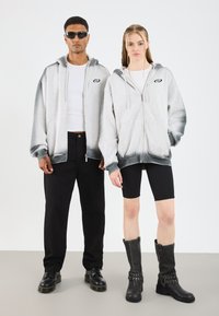 Felpe grigie con zip e cappuccio con sfumature scure nella parte inferiore, pantaloni neri e stivali neri con borchie. Sotto indossato un top bianco semplice.