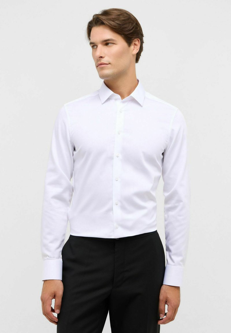 Man in een goed passend wit dresshirt met lange mouwen en zwarte broek, zijkant kijkend tegen een effen achtergrond.