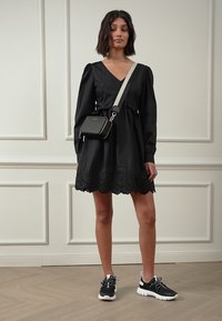 Vestido preto com mangas longas e bainha de renda; usado com uma bolsa de ombro e ténis pretos. Tecido texturizado, corte descontraído.