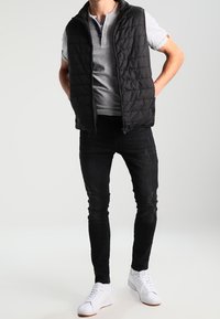 Chaleco acolchado negro sobre una camiseta polo de manga corta gris, combinado con jeans negros desgastados y zapatillas blancas. Fondo neutro.