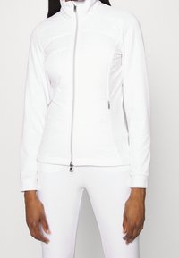 Kobieta ubrana w dopasowaną białą kurtkę z zamkiem błyskawicznym i zamykanymi na zamek kieszeniami z boku oraz pasującą białą spódnicę na jednolitym tle.