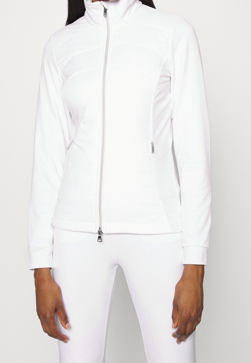 Kobieta ubrana w dopasowaną białą kurtkę z zamkiem błyskawicznym i zamykanymi na zamek kieszeniami z boku oraz pasującą białą spódnicę na jednolitym tle.