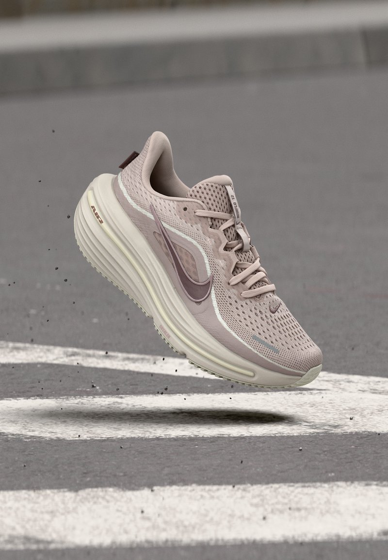 Ανοιχτό μωβ αθλητικό παπούτσι Nike με σχέδιο από δίχτυ και μαλακή σόλα στον αέρα πάνω από άσφαλτο με λευκές οδικές σημάνσεις.