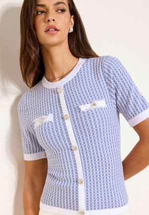 Kvinde iført en kortærmet, lyseblå og hvid mønstret cardigan med knapper foran og to små frynsete lommer.