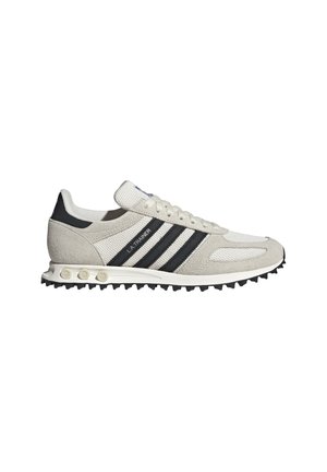 Sneaker Adidas LA Trainer low-top bianca e grigia con strisce nere e suola in gomma testurizzata con dettagli circolari.
