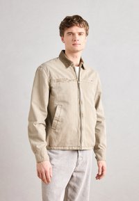 Chaqueta ligera beige con cierre frontal, dos bolsillos laterales y un cuello de contraste. Tejido texturizado y corte casual, combinada con pantalones gris claro.