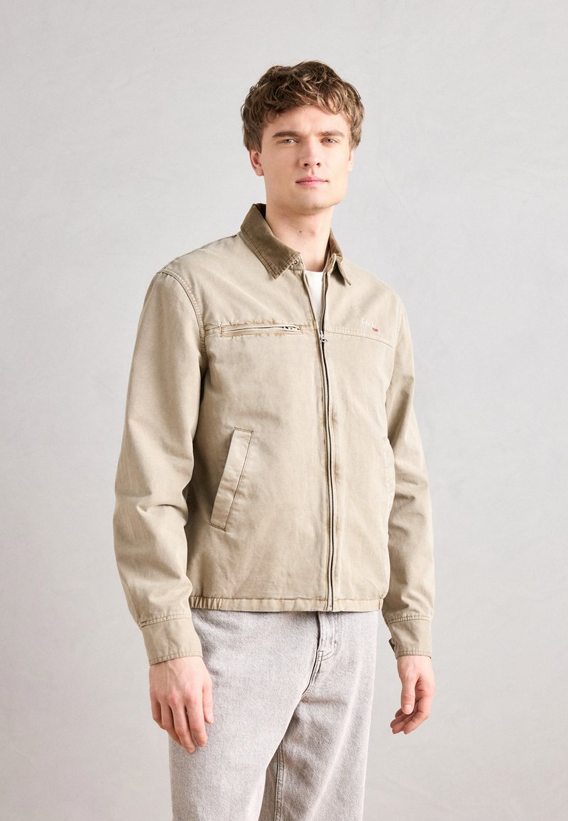 Chaqueta ligera beige con cierre frontal, dos bolsillos laterales y un cuello de contraste. Tejido texturizado y corte casual, combinada con pantalones gris claro.