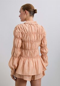 Blusa de manga larga color durazno con detalles de fruncido, cuello con solapa y dobladillo con volantes. Hecha de una tela ligera y texturizada.