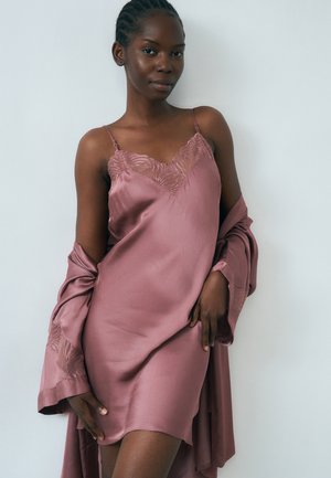 Vrouw draagt een mauve satijnen slipjurk met kanten zoom en een bijpassend ochtendjas over haar armen gedrapeerd, staand tegen een effen lichte achtergrond.