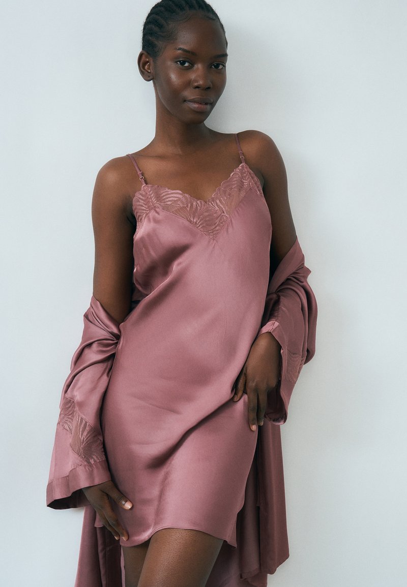 Ženska, ki nosi vijolično satenasto obleko z čipkastim robom in ujemajočim se ogrinjalom, premetenim čez roke, stoji ob preprostem svetlem ozadju.