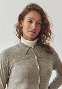 Grauer Strickcardigan mit spitzem Kragen, der eine Knopfleiste hat und darunter einen gerippten Rollkragenpullover trägt. Weicher Stoff und lässige Passform.
