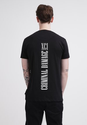Homme tatoué portant un t-shirt noir avec le texte vertical blanc « XCI » et « CRIMINAL DAMAGE » au dos, sur un fond uni.