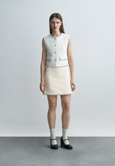 Jeune femme posant devant un fond uni, vêtue d'un gilet blanc texturé sans manches, d'une jupe crème avec bordure décorative, de chaussettes claires et de talons noirs.