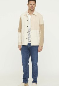 Lichte, bicolore jas in crème en beige, met een relaxte pasvorm, zijzakken en zwarte knopen, over een bloempatronen shirt en spijkerbroek.