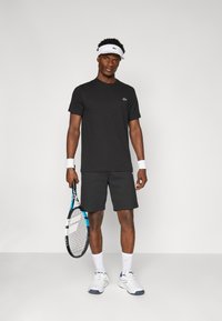 Zwarte sport T-shirt en shorts, witte tennisschoenen, dragend een witte visor en polsbandjes, met een tennisracket met blauwe accenten.