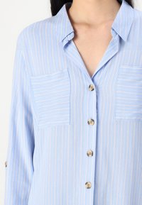 Light blue striped shirt, finished with a collar, two chest pockets, and buttons down the front. Soft fabric with a smooth texture.