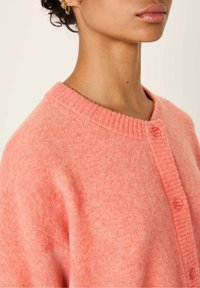 Roza roza pletena jakna z rebrastim okroglim izrezom, zapenjanjem na gumbe s kroglastimi gumbi in mehko teksturo.