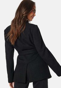Sort blazer med en tekstureret stof, lange ærmer og en bæltet talje. Har en bagåbning for nem bevægelse. Ren, skræddersyet silhuet.