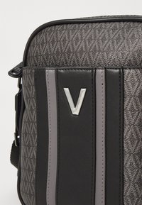 Gros plan sur un sac texturé noir et gris avec des rayures verticales et une lettre "V" métallique centrée sur le panneau avant.