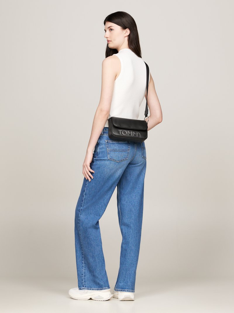 Bolsa de hombro de cuero negro con el logo "TOMMY" en relieve, llevada sobre una blusa blanca sin mangas y unos jeans rectos de pierna ligera en azul, combinada con zapatillas blancas.